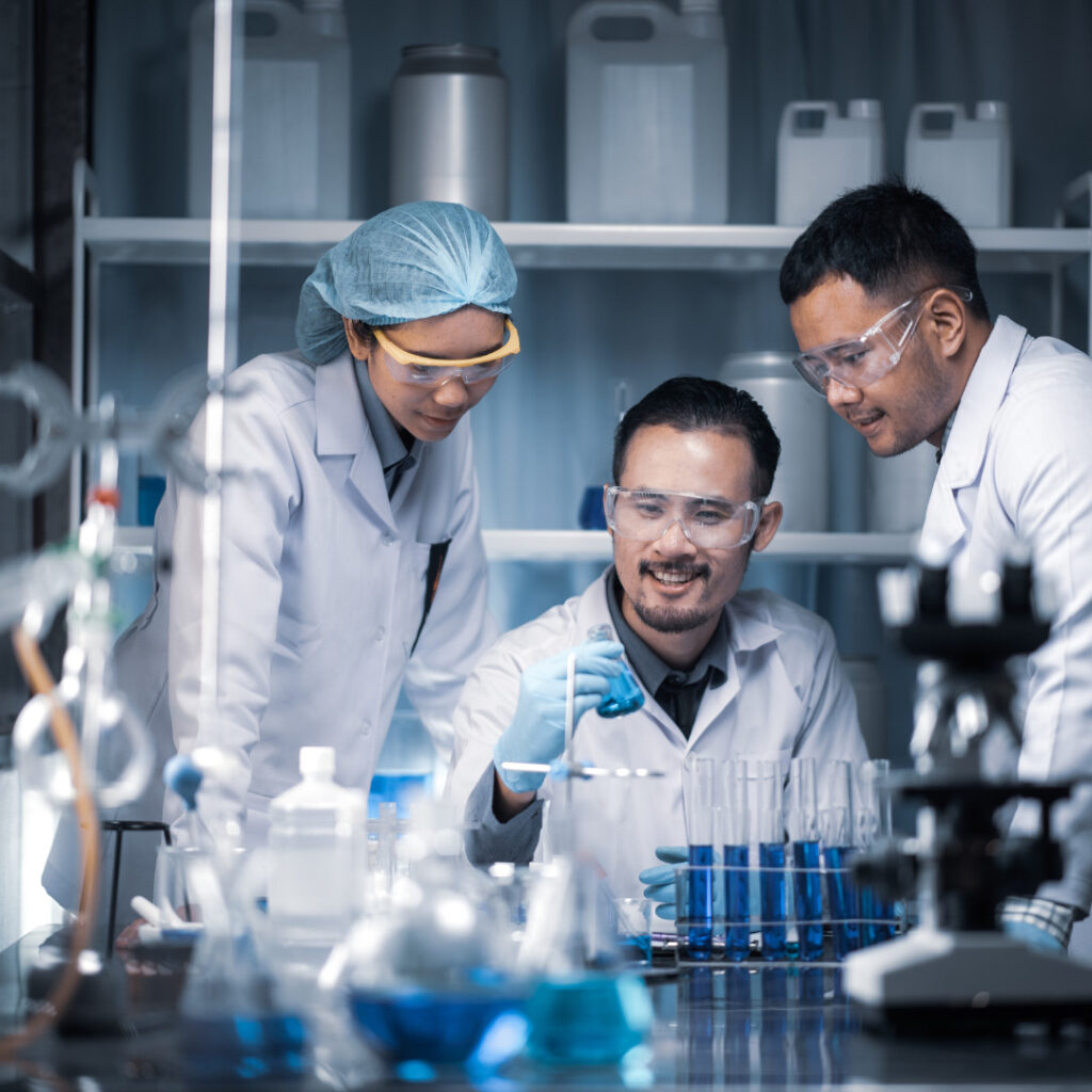 Three researchers in lab coats and safety goggles collaborate at a lab bench, examining blue liquid in beakers and test tubes