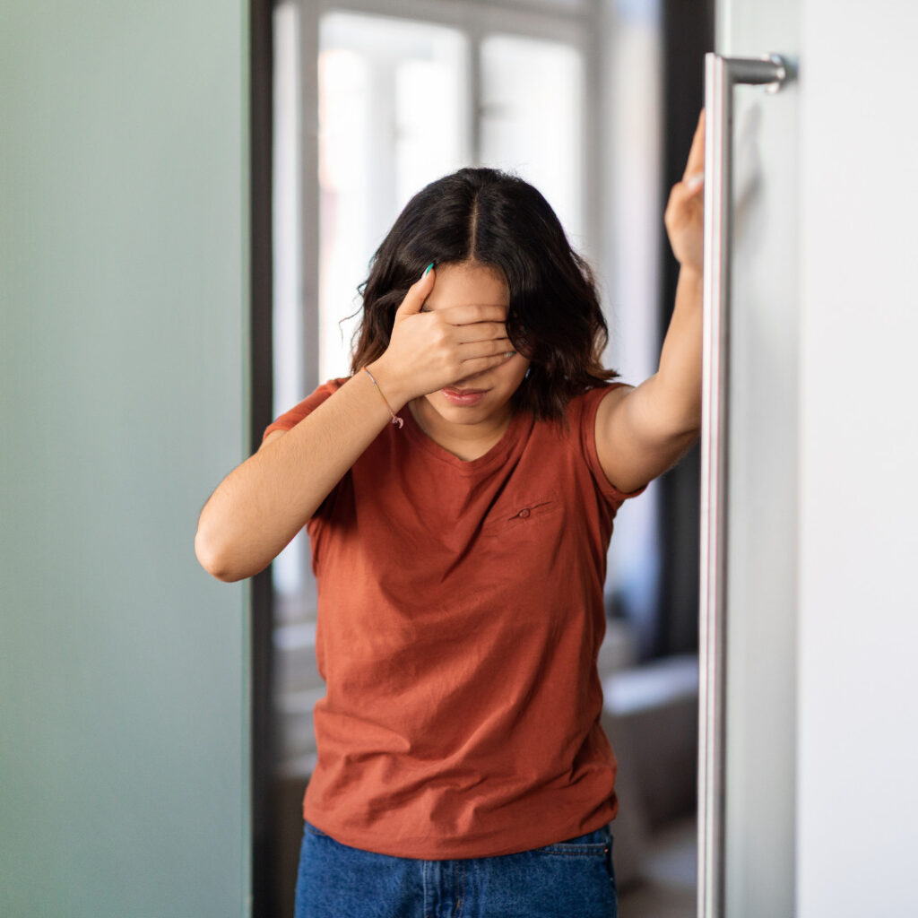 A young woman standing in a doorway, bracing herself against the door frame with one hand while covering her eyes with the other, suggesting dizziness or lightheadedness upon standing.
