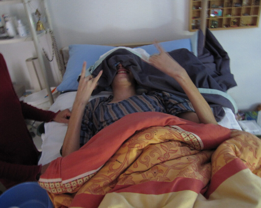 Jonas lies in bed wearing an eye mask while a loved one holds his hand — illustrating daily life with severe ME/CFS.