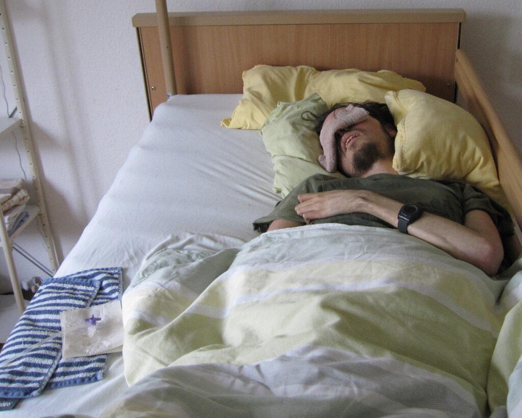 Jonas resting in his care bed with a pink eye mask, surrounded by soft yellow and green bedding.