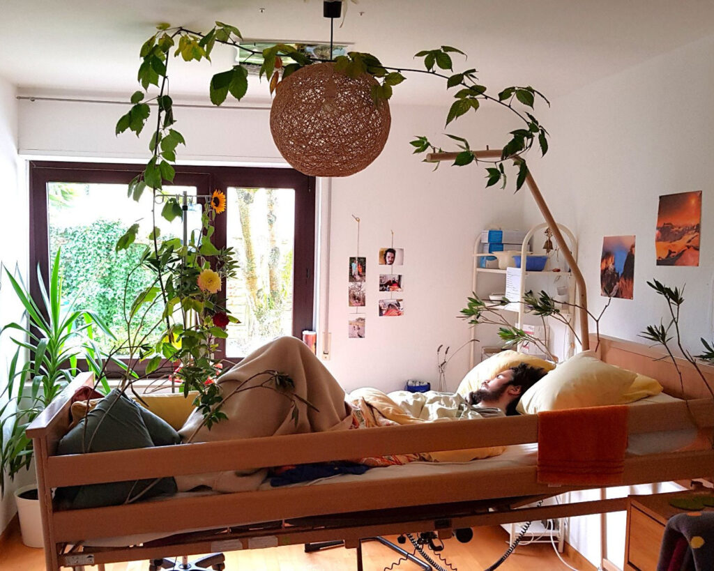 Jonas in his bedroom surrounded by sunflowers, hanging plants, and family photos on the wall.