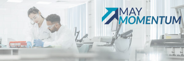 Two researchers in white lab coats and safety glasses working with samples in a bright laboratory, with the blue "May Momentum" logo overlaid upper right.