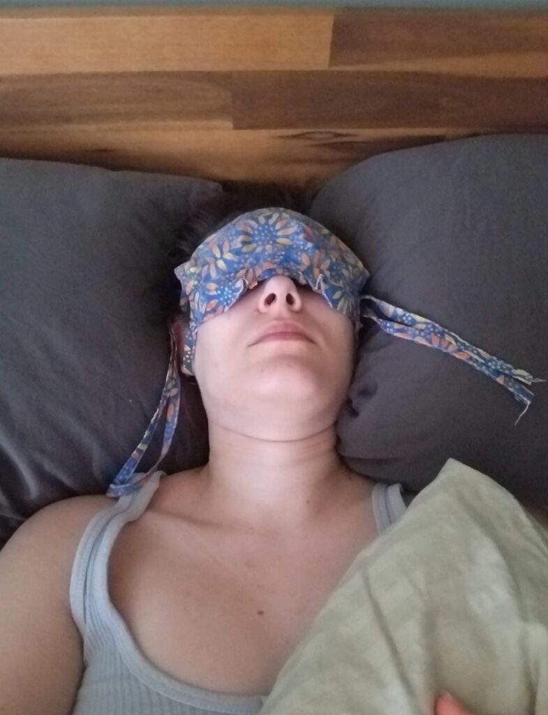 Tina lying in bed wearing a floral eye mask to block light, illustrating life with ME/CFS.