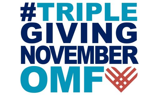 TGN logo: #TRIPLEGIVINGNOVEMBEROMF. The phrase is written in bold, uppercase letters with a mix of dark blue and light blue colors. The "OMF" text is in light blue, and there is a stylized heart shape in reddish-orange next to it.